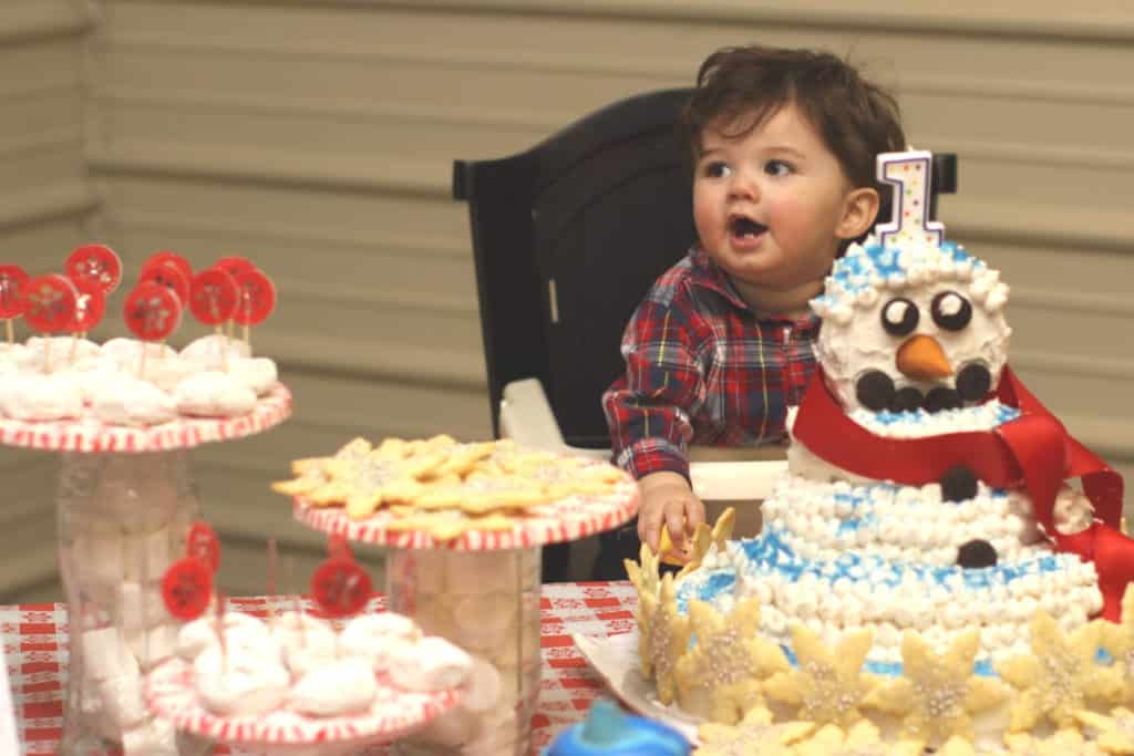 Baby sitting at a festive snowman themed birthday party with goodies and a snowman birthday cake