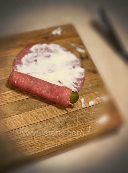 Cutting board on a kitchen counter with dill pickle getting rolled up inside Buddings dried beef luncheon meat and cream cheese to make dill pickle roll up recipe.