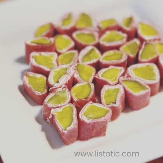 Green dill pickles wrapped in cream cheese and dried beef on a white appetizer plate.