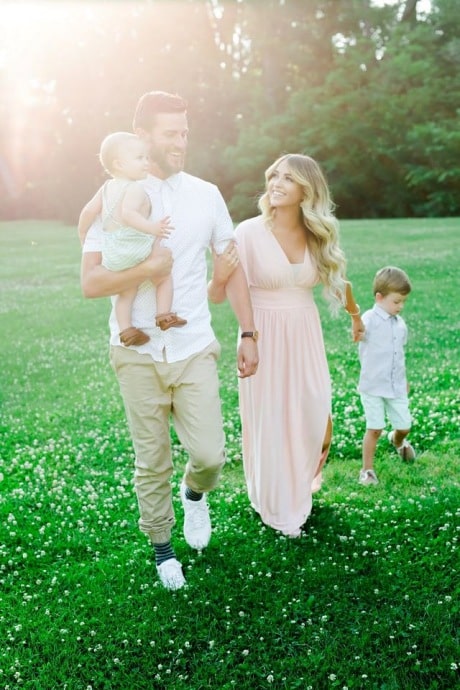 family of four holding hands in the summer time wearing neutral beach colors in their family photo outfit. Mom smiling at dad holding a baby and walking together in green grass. 