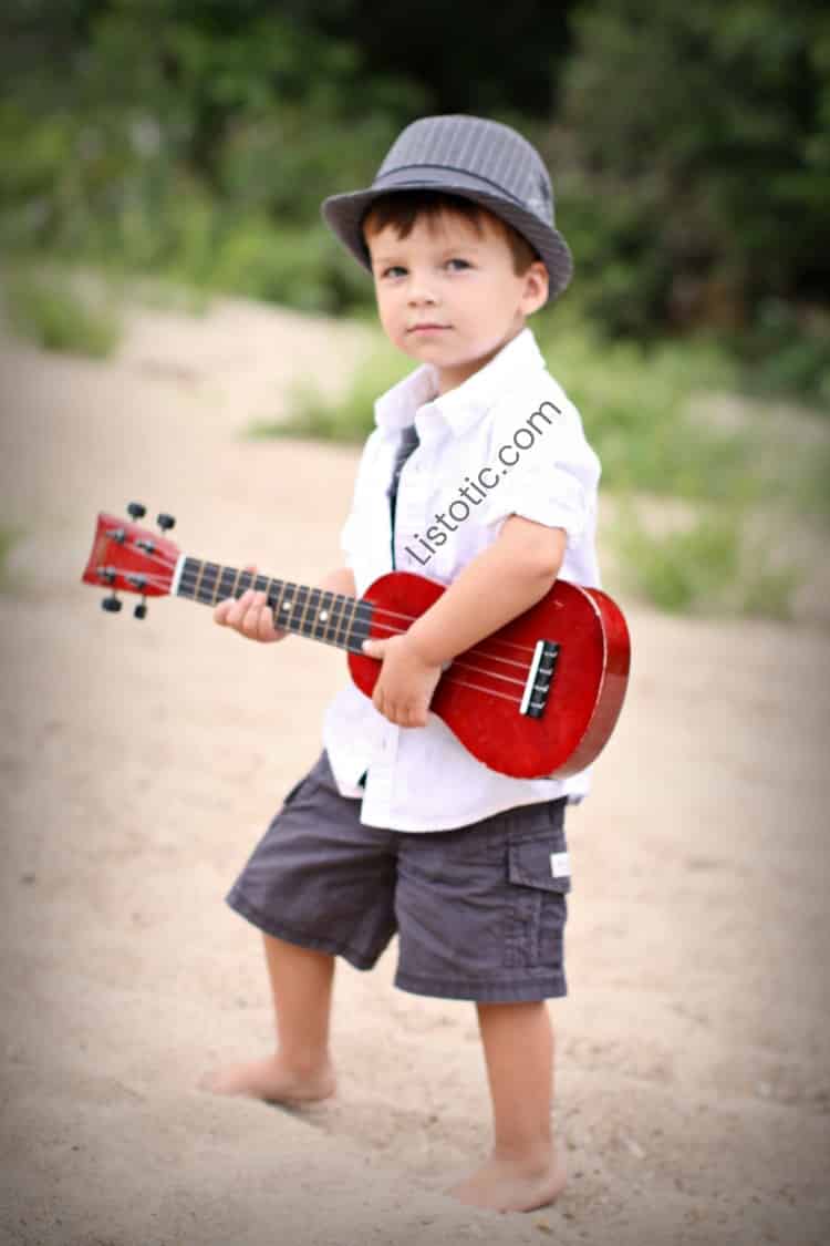 boy wearing a hat standing on a beach and holding a small red guitar 