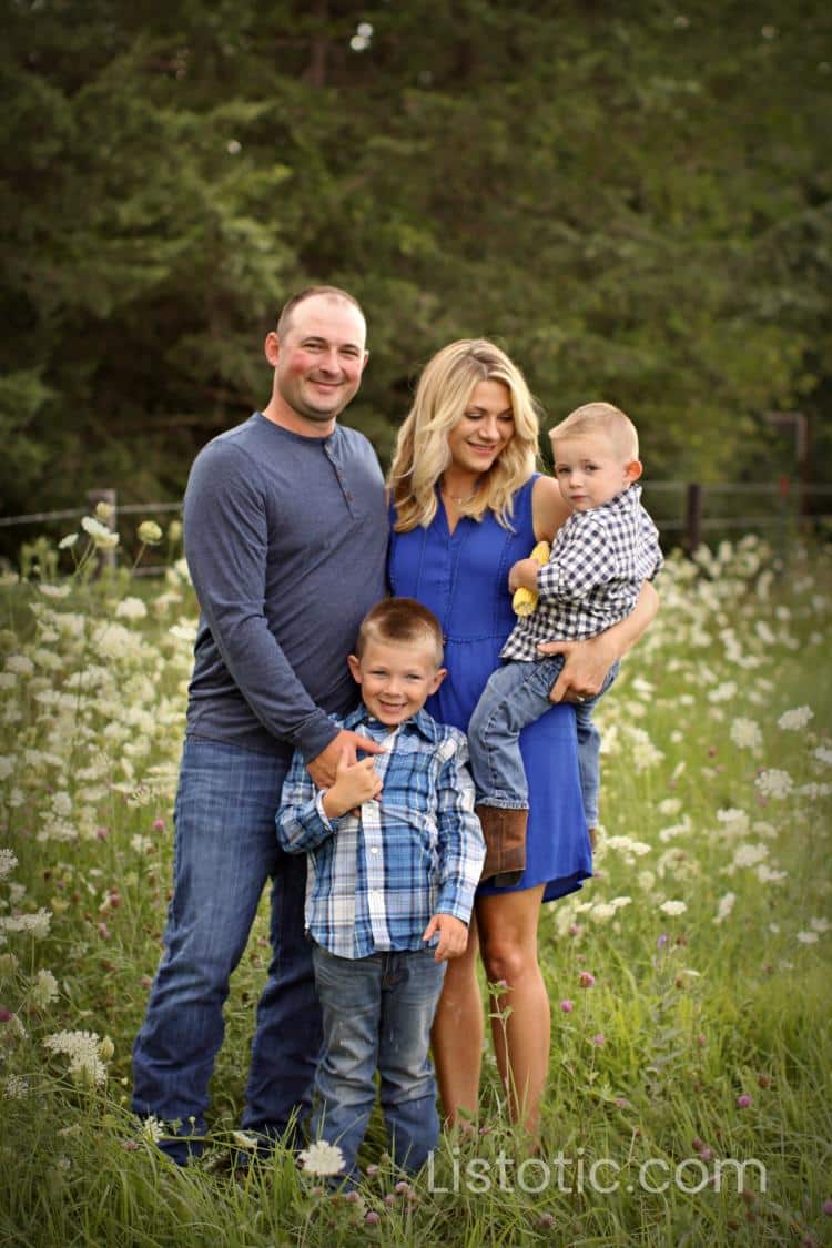 family of four wearing different shades of blue standing in a tall grass field. Mother of two boys smiling at baby boy. Father proudly holding son’s shoulder in posed family portrait. 