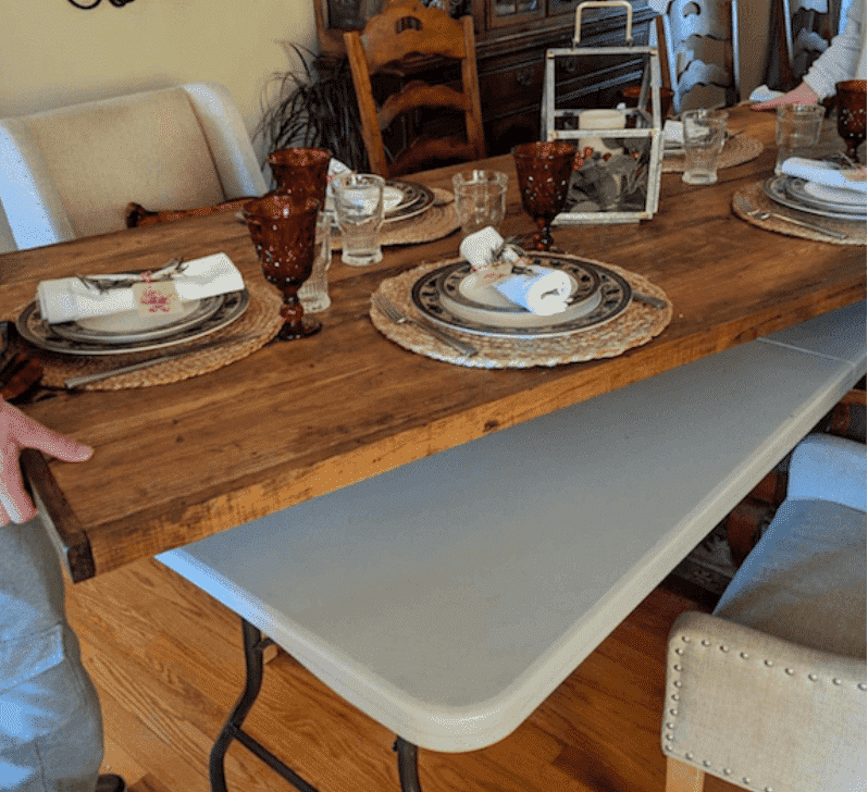 Wooden table topper set with fancy glasses and plates being placed by two people holding it on either side on top of a plastic folding table in a home for thanksgiving dinner.