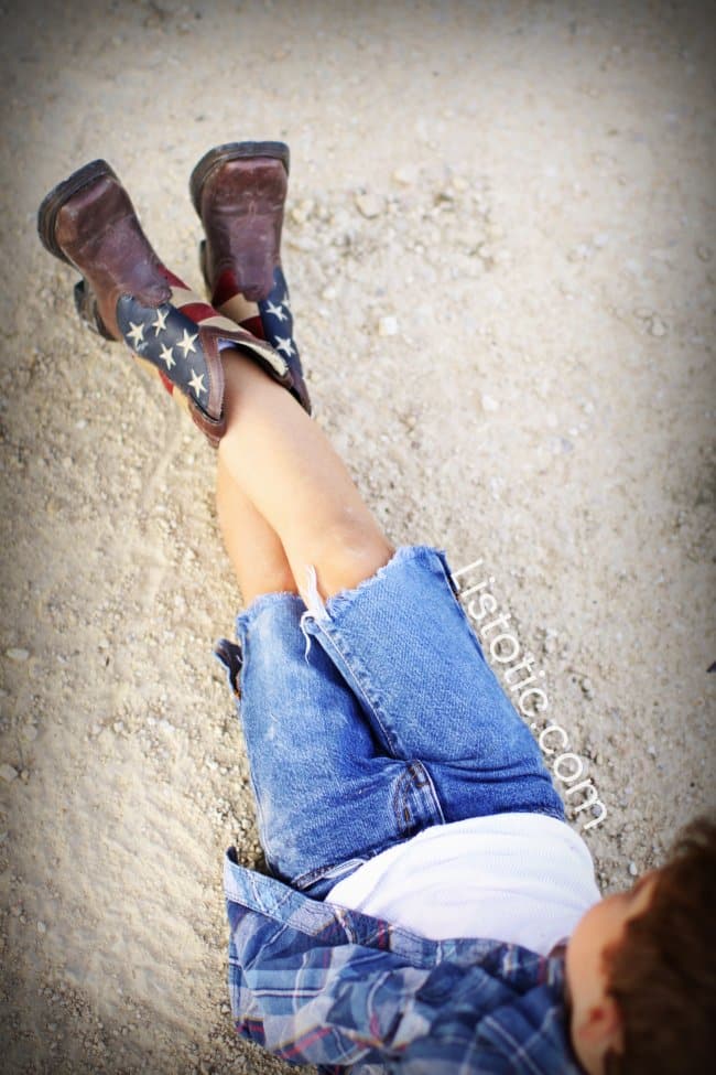 Photo from above of a boy sitting on gravel road with legs stretched out. Wearing cowboy boots, cut off jean shorts and flannel shirt. 