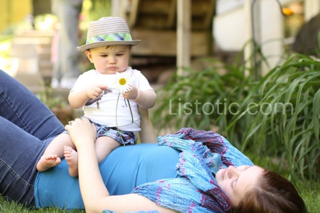 woman laying on back on the grass wearing a blue scarf and a baby sitting her stomach wearing a straw hat and holding a daisy. 