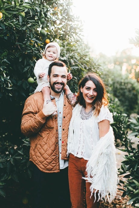 Happy family of 3 in neutral colors and layered jackets and vests. Baby on the shoulders of dad standing together and smiling in front of an orange tree. 