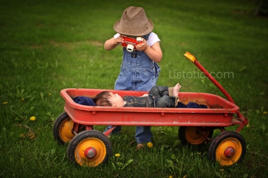 toddler in bib overalls and hat holding a Fischer price play camera leaning over a baby laying on his back in red wagon also wearing bib overalls. Boy standing is pretending to take a photo of the baby in the wagon. 