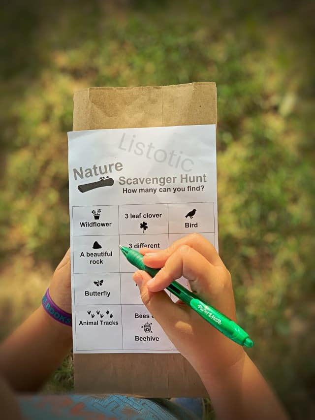 brown paper sack with free scavenger hunt printable attached. Child using a green pen to mark of the collected scavenger hunt items in a backyard or park. 