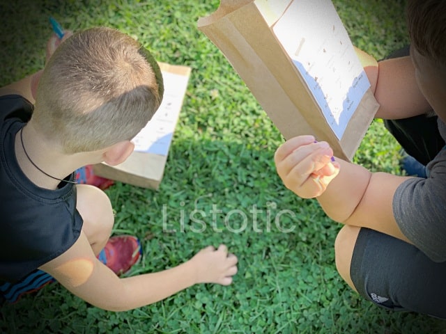 two kids looking for clues on their nature scavenger hunt brown paper sacks with a bingo type design for a nature scavenger hunt outside in the park or backyard. 