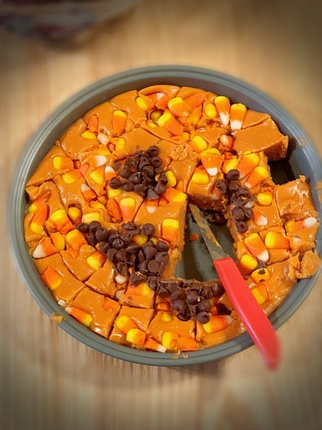 Cake pan of candy corn butterfinger fudge cut with knife still in it and pieces missing from the center of the dessert. 