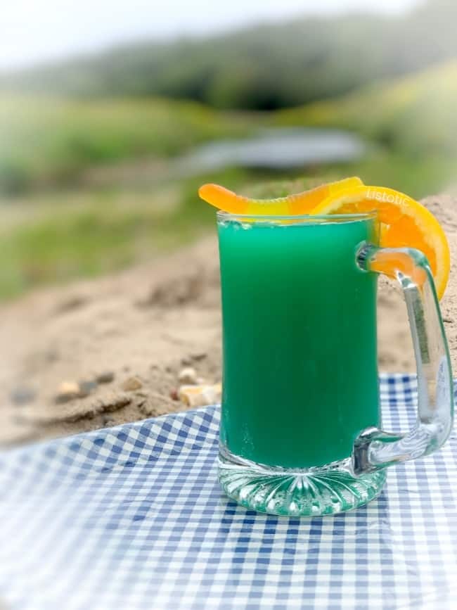 A greenish blue colored cocktail in a beer mug on a sandy creek side beach. Glass mug garnished with a gummy worm and orange slice on a blue gingham patterned tray.