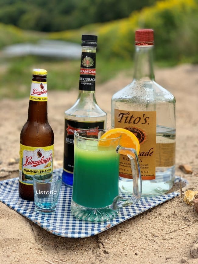 Ingredients to create a river water beer cocktail recipe. Summer shanty beer, blue curacao orange liquor, vodka liquor, orange juice and a slice of orange. Ingredients resting on a gingham patterned metal tray next to a slow creek in the country side. 