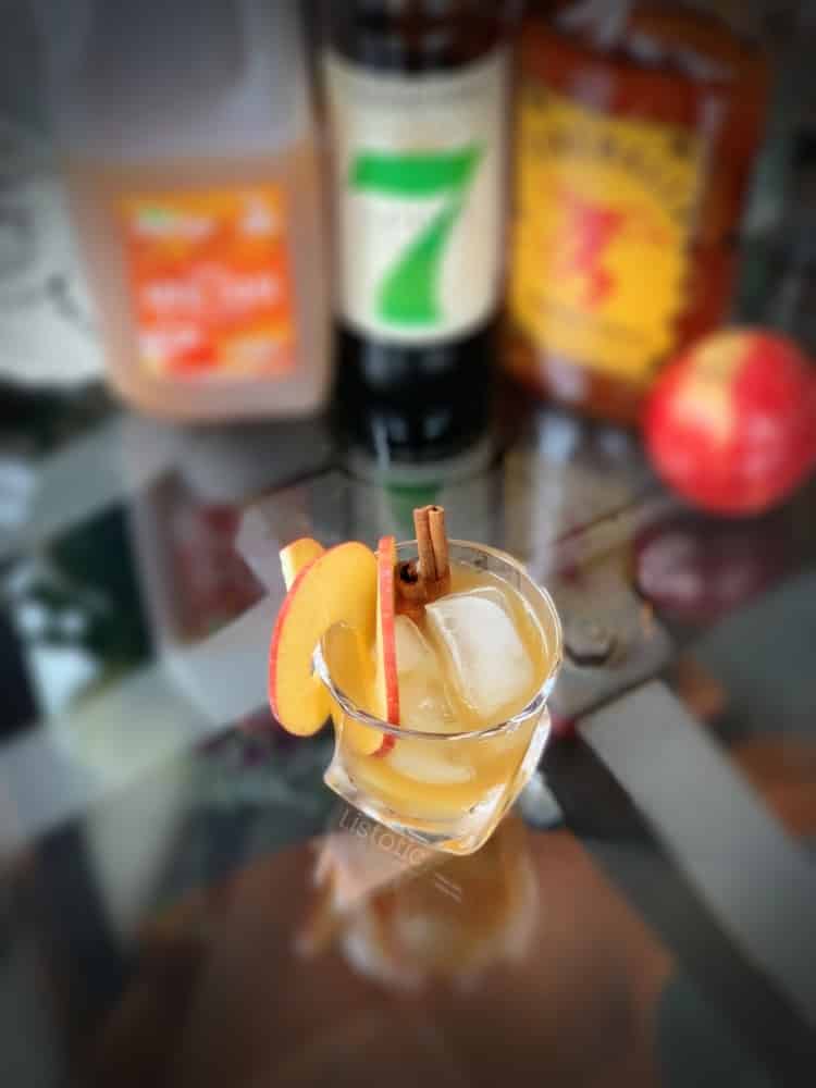 Looking down into a glass of apple whiskey with fireball and apple cider. Glass has garnish of apple slices and cinnamon sticks. Jug of apple cider and liquor bottles sitting on the table nearby.