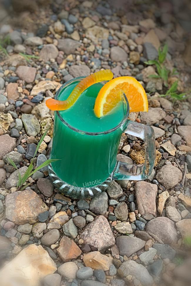 Beer mug full of blueish green river water cocktail recipe on a rocky river bank garnished with orange slice and gummy worm. 