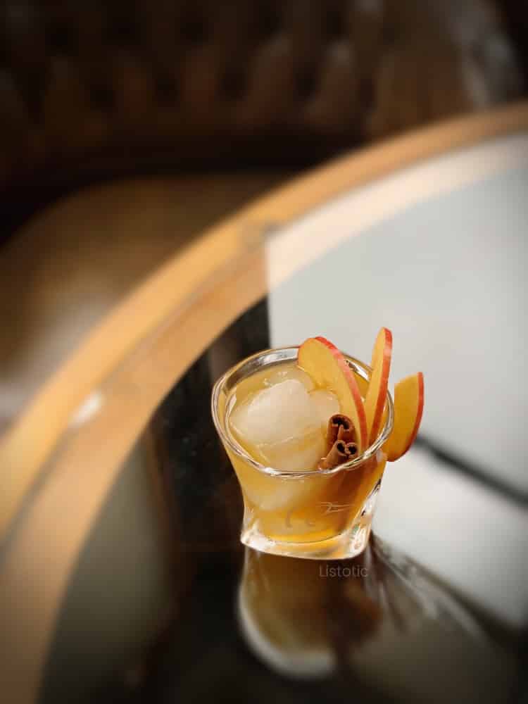 Fancy short glass filled with ice, whiskey and apple cider sitting on a glass top table next to a window. Cocktail glass is garnished with a spiral slice of apple and cinnamon sticks. 