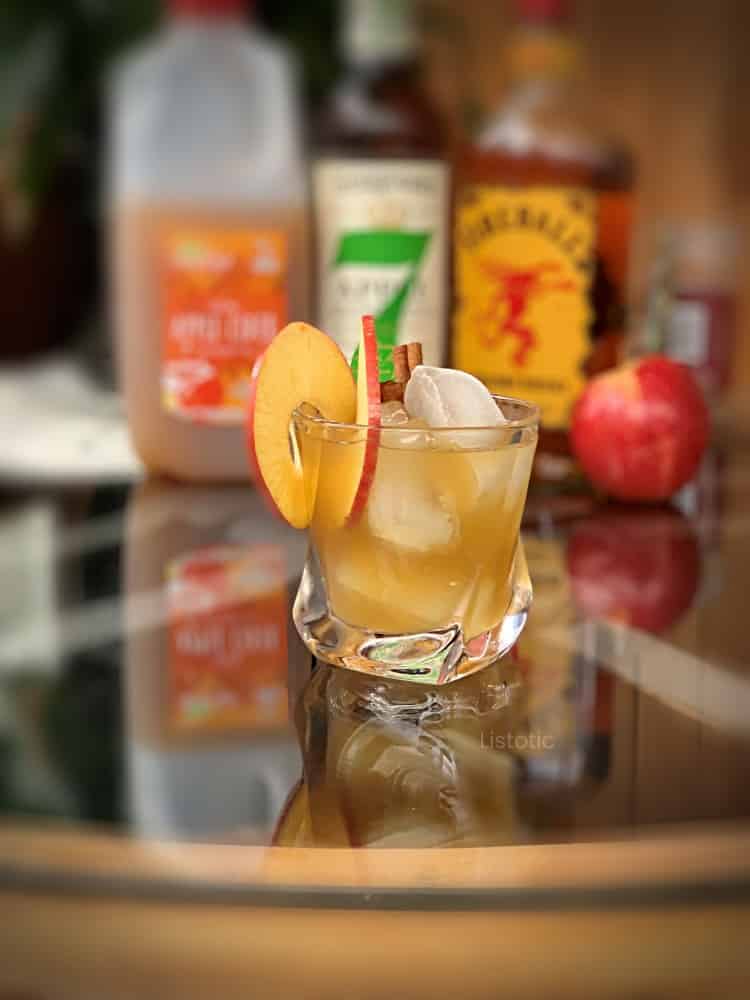 Clear cocktail glass filled with apple cider, liquor and ice. Spiral sliced apple garnish and whiskey cinnamon apple cocktail ingredients in the background. 