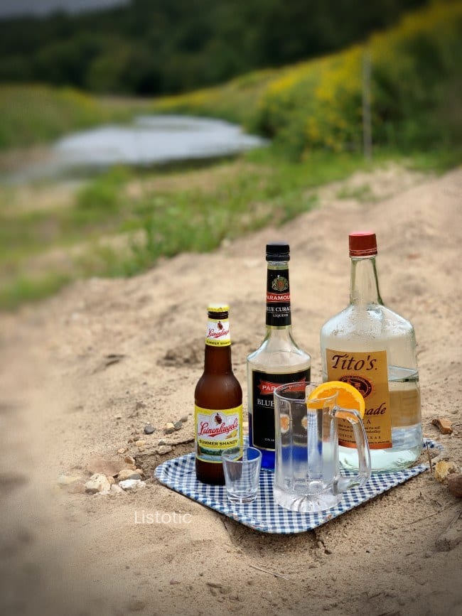 Ingredients for a beer cocktail recipe sitting on a metal tray near a sandy bank of a beautiful country side creek on a summer day. 