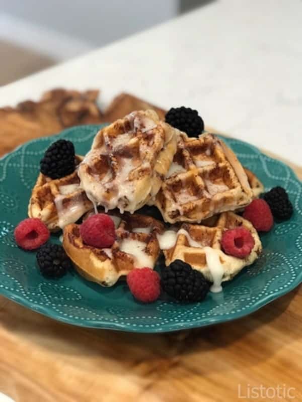 A blue plate with stacked cinnamon roll waffles drizzled in melted cream cheese frosting topped with fresh berries.