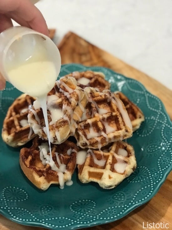 Stacked Cinnamon Roll Waffles on a blue lace decorative plate being drizzled with melted cream cheese frosting. 