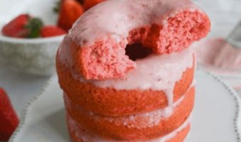 Four glazed strawberry donuts stacked on a white scalloped cake stand with a white bowl filled with strawberries.