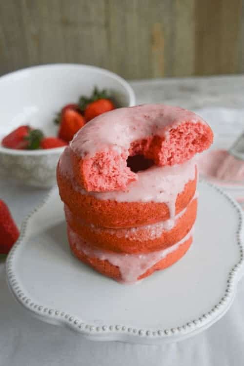 Four glazed strawberry donuts stacked on a white scalloped cake stand with a white bowl filled with strawberries.