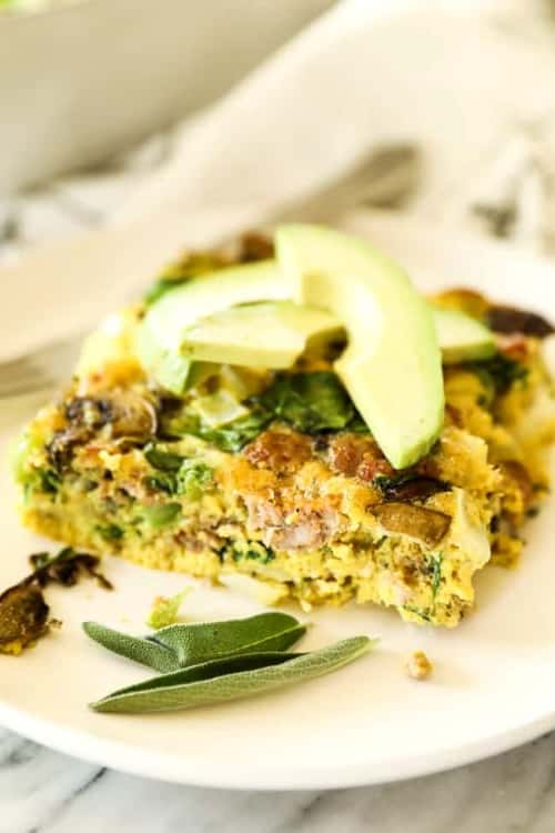 Keto friendly egg breakfast casserole slice topped with sliced avocado and sage leaves.