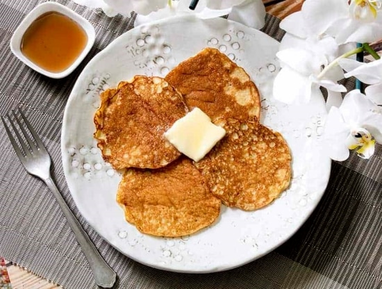 Four Small browned pancakes on a white plate topped with melting butter and a side of syrup.