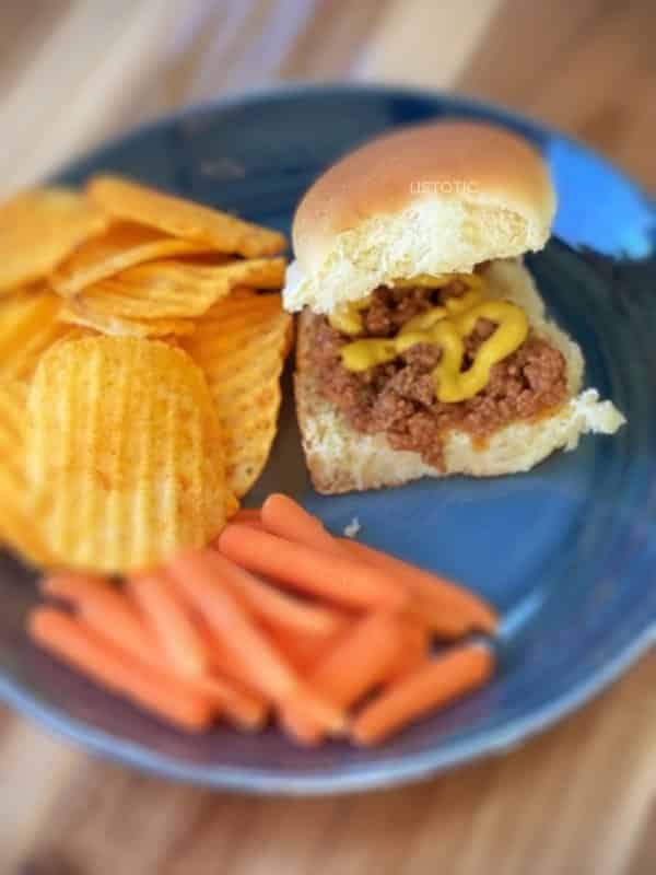 Loose meat sandwich with mustard on a blue plate with sides of carrots and chips