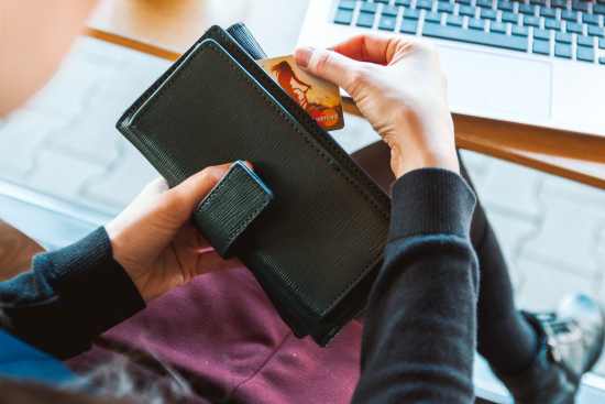 A person holding a black wallet with credit card and lap top in the background.