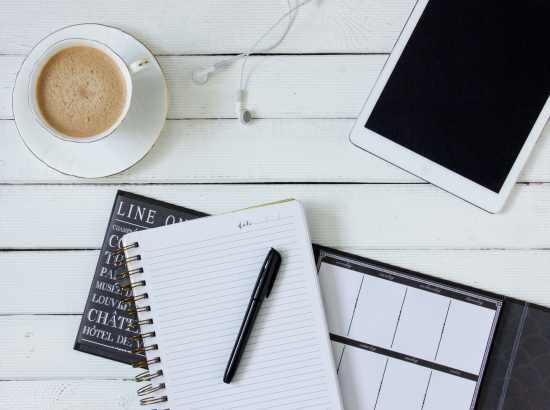 A table with an ipad, calendar, notepad with pen and a cup of coffee ready to plan.