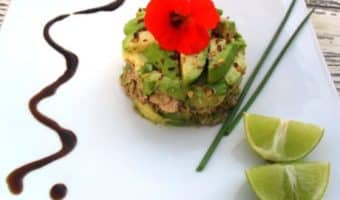 An avocado tartare displayed on a plate with chopsticks, lime slices, and a drizzle of sauce on the side.