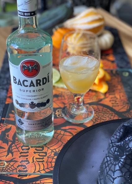 Bacardi White Rum bootle next to a glass of orange juice and witches hat to make zombie cocktail drink