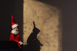 Christmas elf is back and he is sitting on the corner of the tv with his shadow showing on the wall behind him from the morning sun